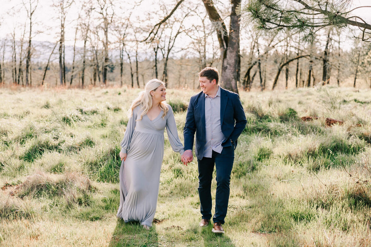Portrait of a husband and wife walking holding hands smiling at each other. She is wearing a silver gown and he is wearing a suit jacket and slacks.
