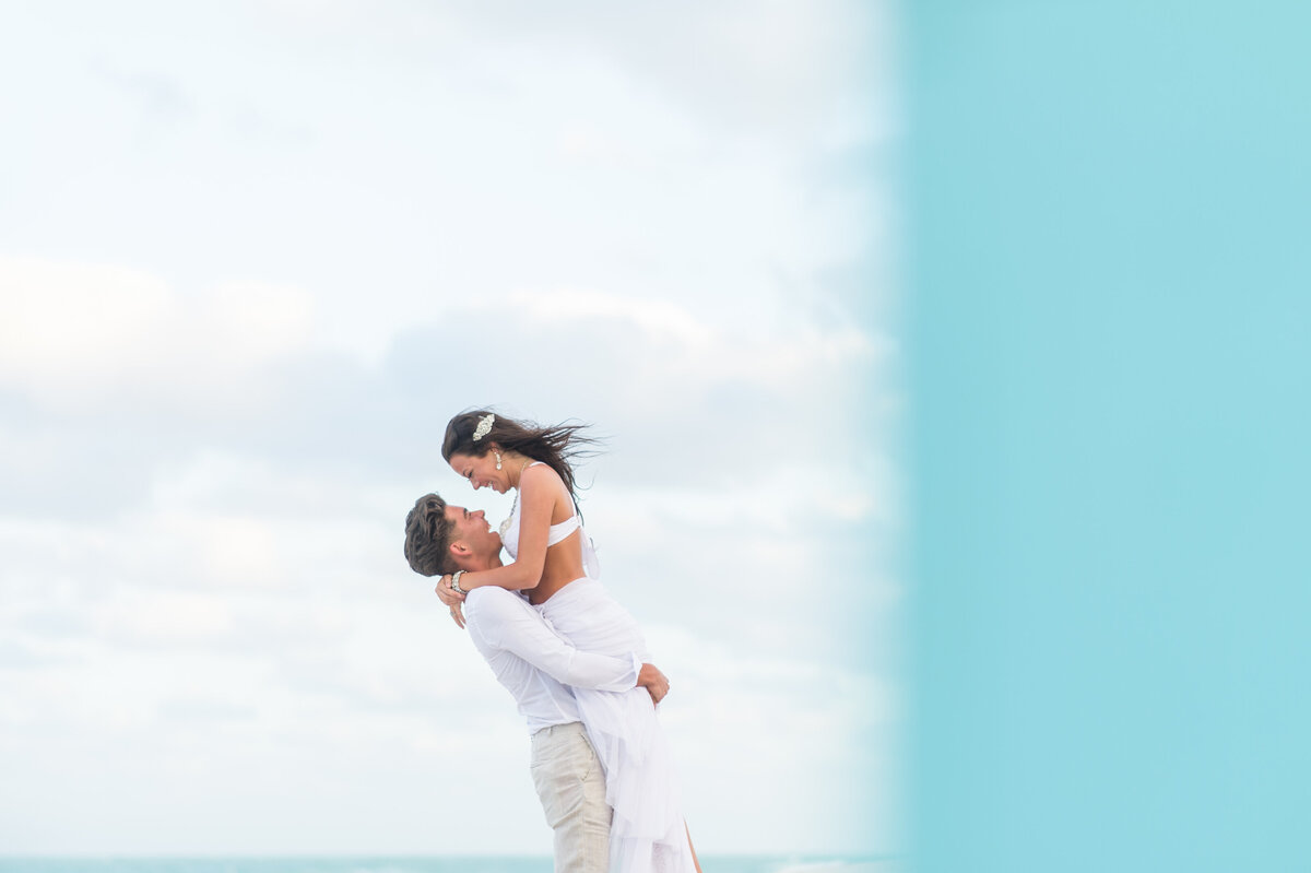 Romantic couple sharing a joyful moment on a Miami beach as the man lifts the woman against a backdrop of soft clouds and ocean.