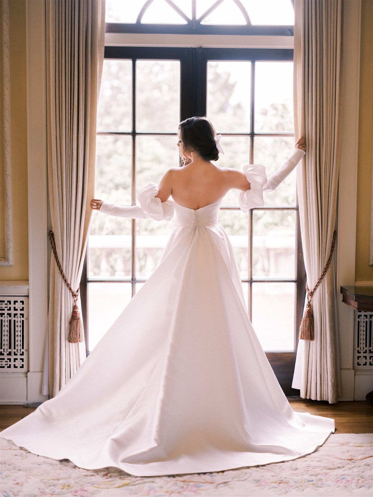 A woman in a strapless white wedding dress stands with her back to the camera, holding open curtain drapes and looking out a large window, curated to perfection by Destination Wedding Planner Melissa Dawn Event Designs.