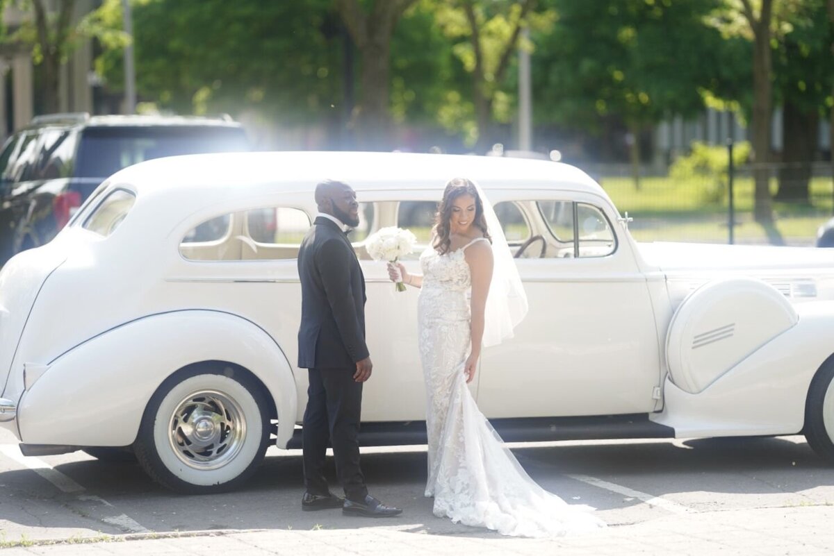 The couple stands together in front of a classic vintage limousine, exuding elegance and sophistication. The bride and groom are dressed in their wedding attire, with the luxurious car serving as a stylish backdrop. The image captures a timeless moment, blending romance with the grandeur of the vintage vehicle.