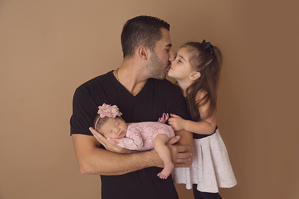 A father in a black shirt kisses his toddler daughter while holding his sleeping newborn baby in his arms in a NJ Newborn Photography studio