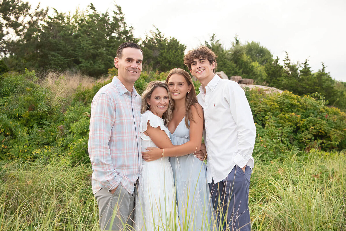 A family poses in the tall grass of Wingaersheek Beach with the mom and daughter with their arms around each other and the dad and son on either end.