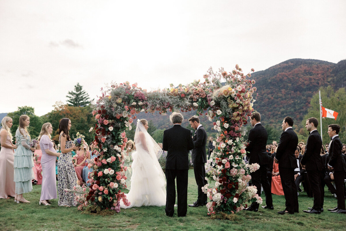 adirondack-mountain-wedding-26-bride-and-groom-under-floral-arch-with-guests-seated-in-garden-chairs-at-adirondack-mountain-wedding-jenny-fu-studio