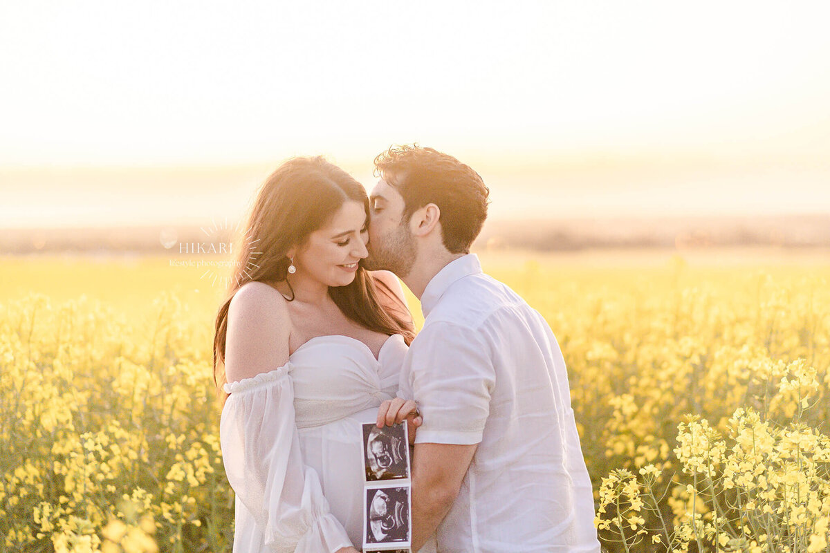 girl holding ultrasound pictures while boy kisses her in flower field near adelaide gold coast