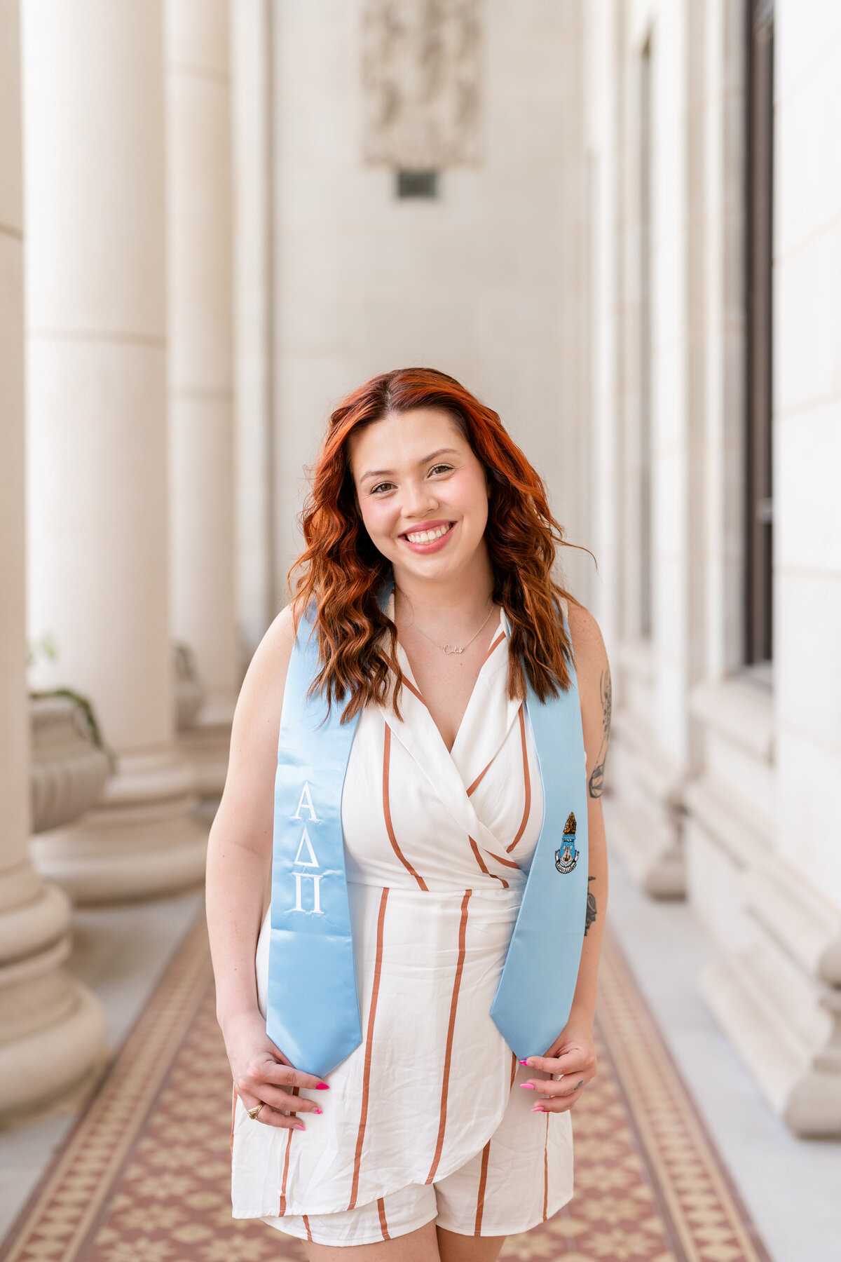 Texas A&M senior girl smiling and holding Pi Phi stole while wearing white stripe romper in the columns of the Administration Building