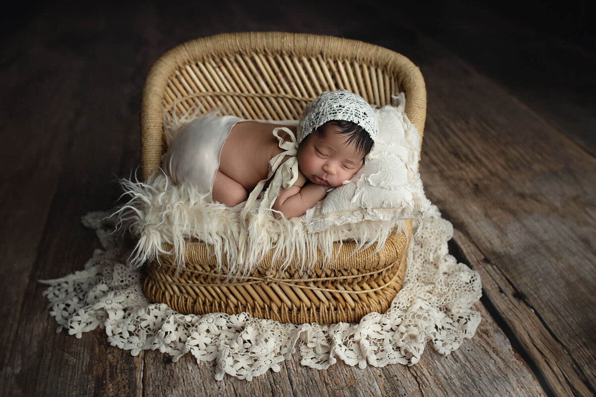 A newborn baby sleeps in froggy pose in a small bonnet on a wicker chair on a lace blanket in a New Orleans Newborn Photographer studio