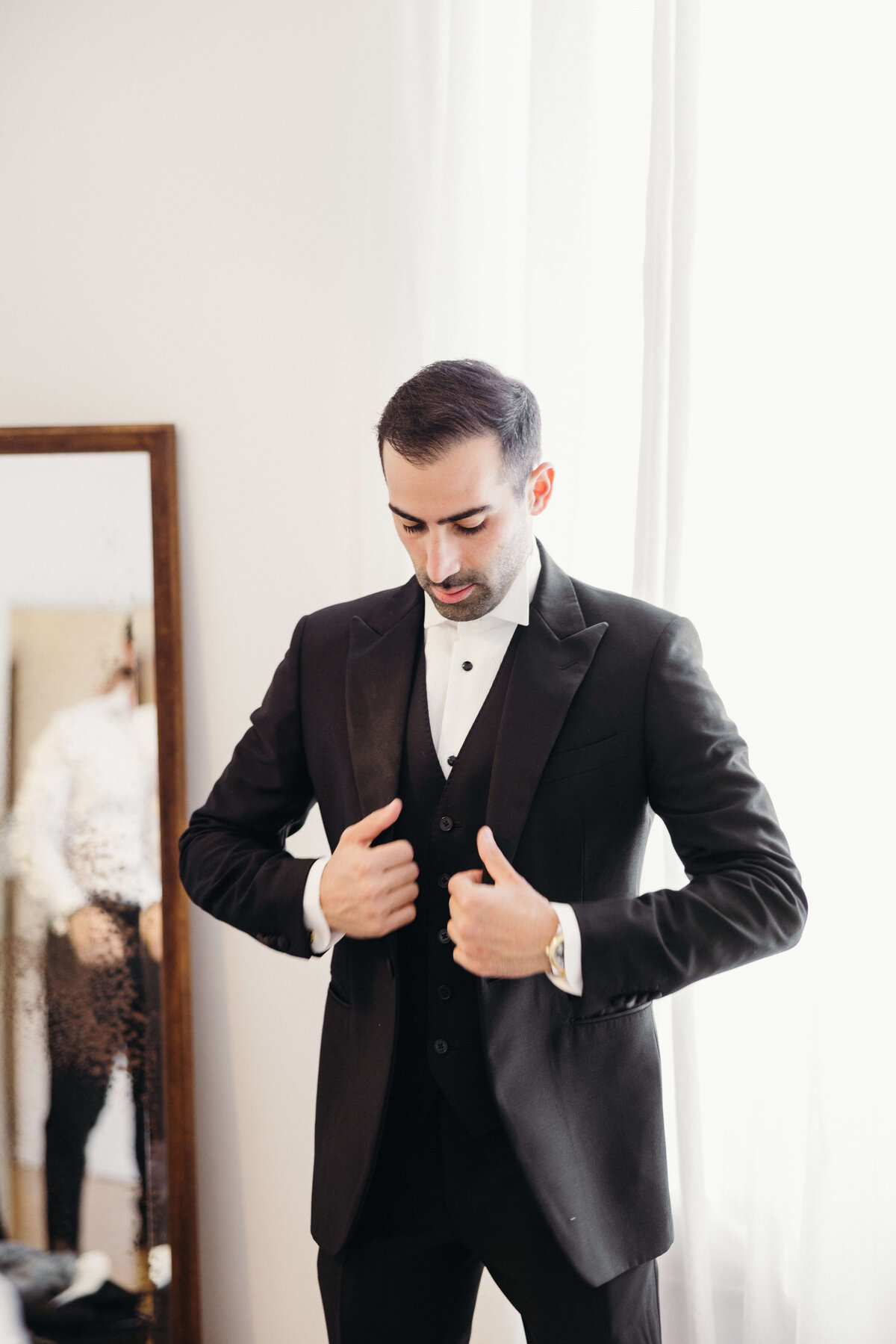 Groom putting on classic black tuxedo jacket in front of mirror

