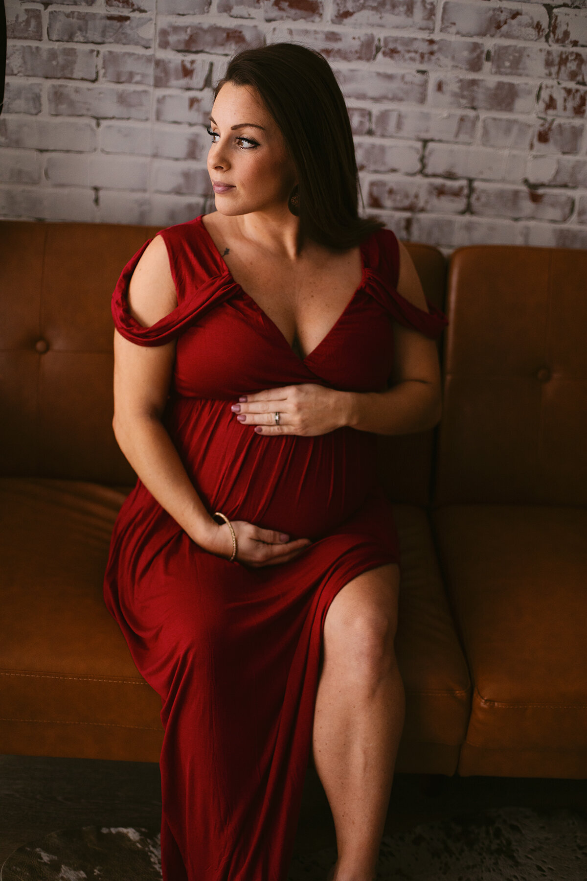 A pregnant woman in a red dress sits on a tan couch while looking out the window during her Bentonville maternity boudoir photoshoot.