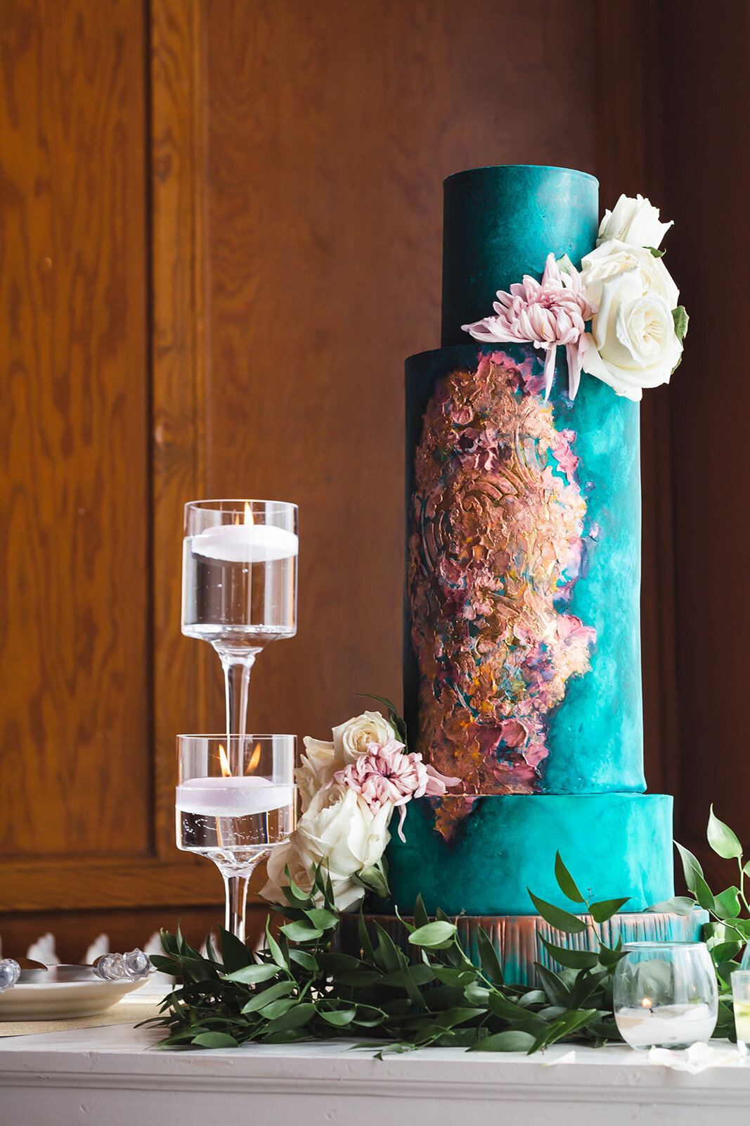 a teal wedding cake beside clear candle holders surrounded by greenery