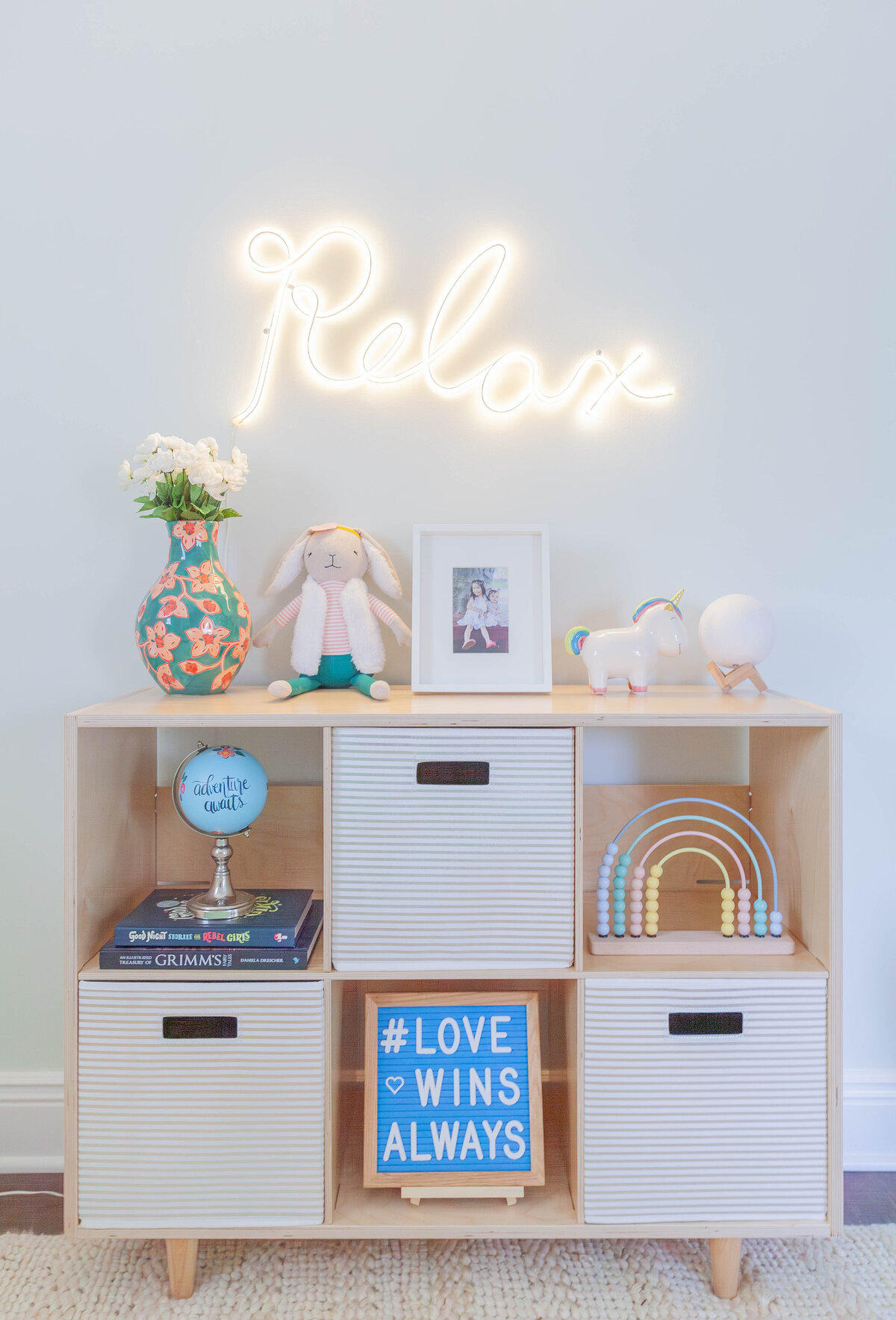 A playful and modern kids’ room vignette by Deni + Dove Interiors featuring a light wood cubby shelf with striped storage bins, children’s books, toys, and a neon 'Relax' sign above. Styled in a Montclair, NJ family home.