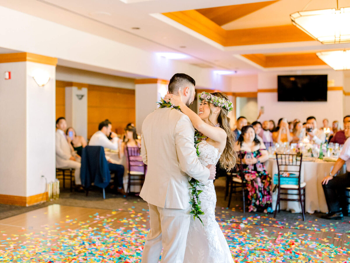 A couple in elegant attire shares a joyful dance at a wedding, surrounded by guests seated at round tables. Colorful confetti scatters the floor, creating a festive atmosphere.