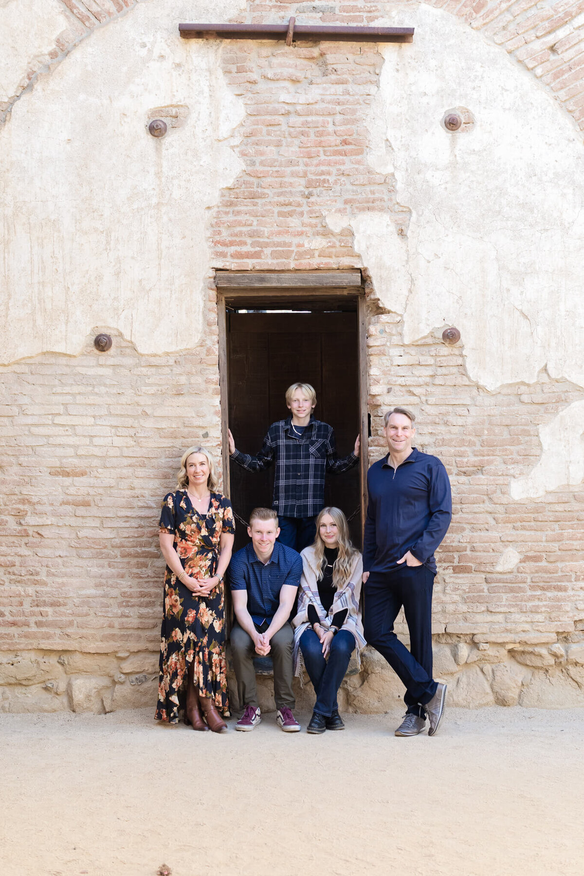 A family of five standing and sitting around a wooden archway against a beige brick wall, looking at the camera.