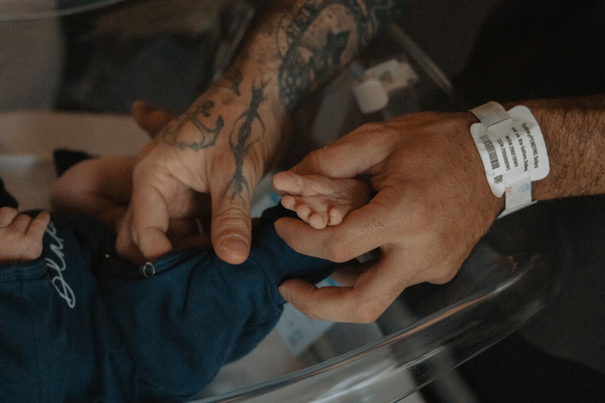 Hands gently holding a newborn's tiny foot, a moment captured with delicate tattoos visible