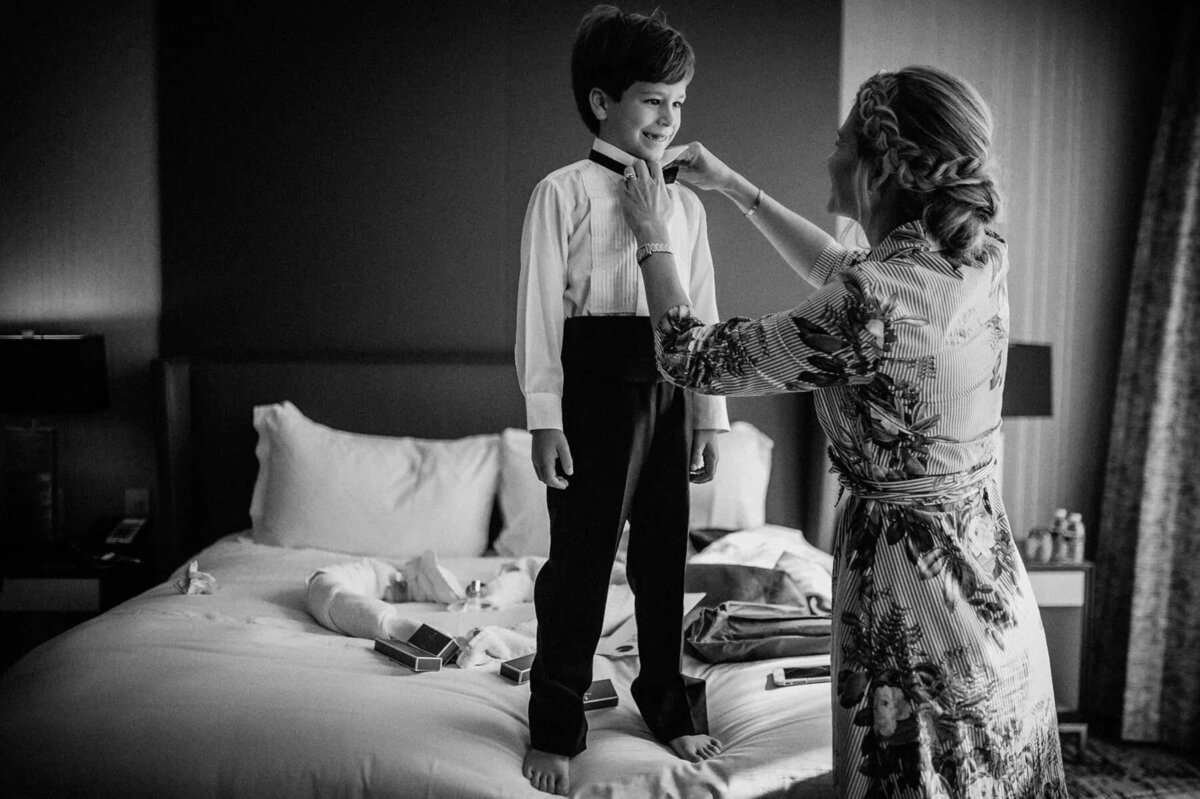 A boy standing on a bed while a woman ties a bowtie on him.