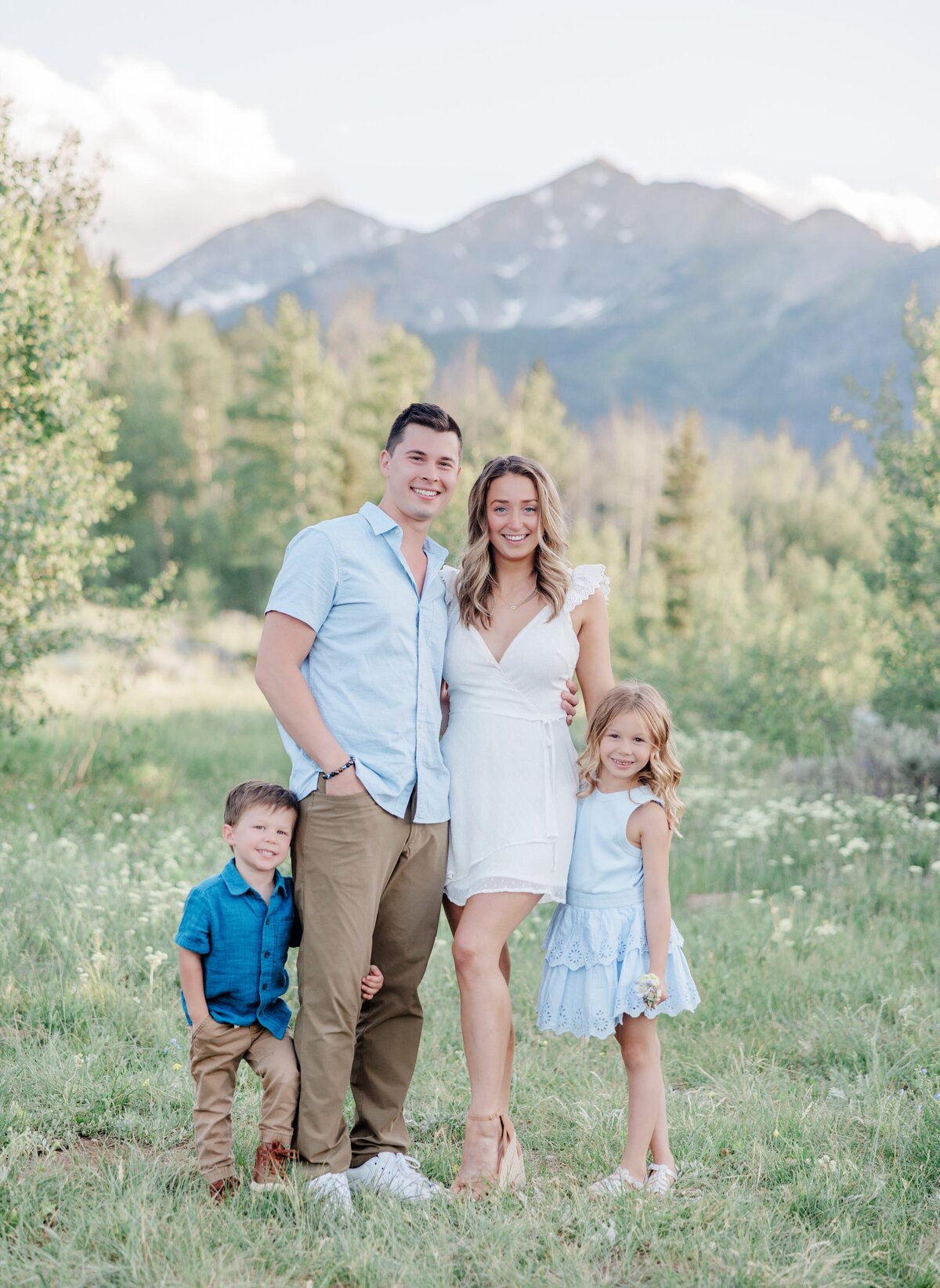 A mom and dad stand in a mountain meadow with their toddler son and daughter hugging their legs