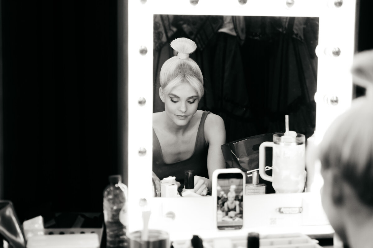 Performer in blonde pruik maakt zich op voor een belichte theaterspiegel.