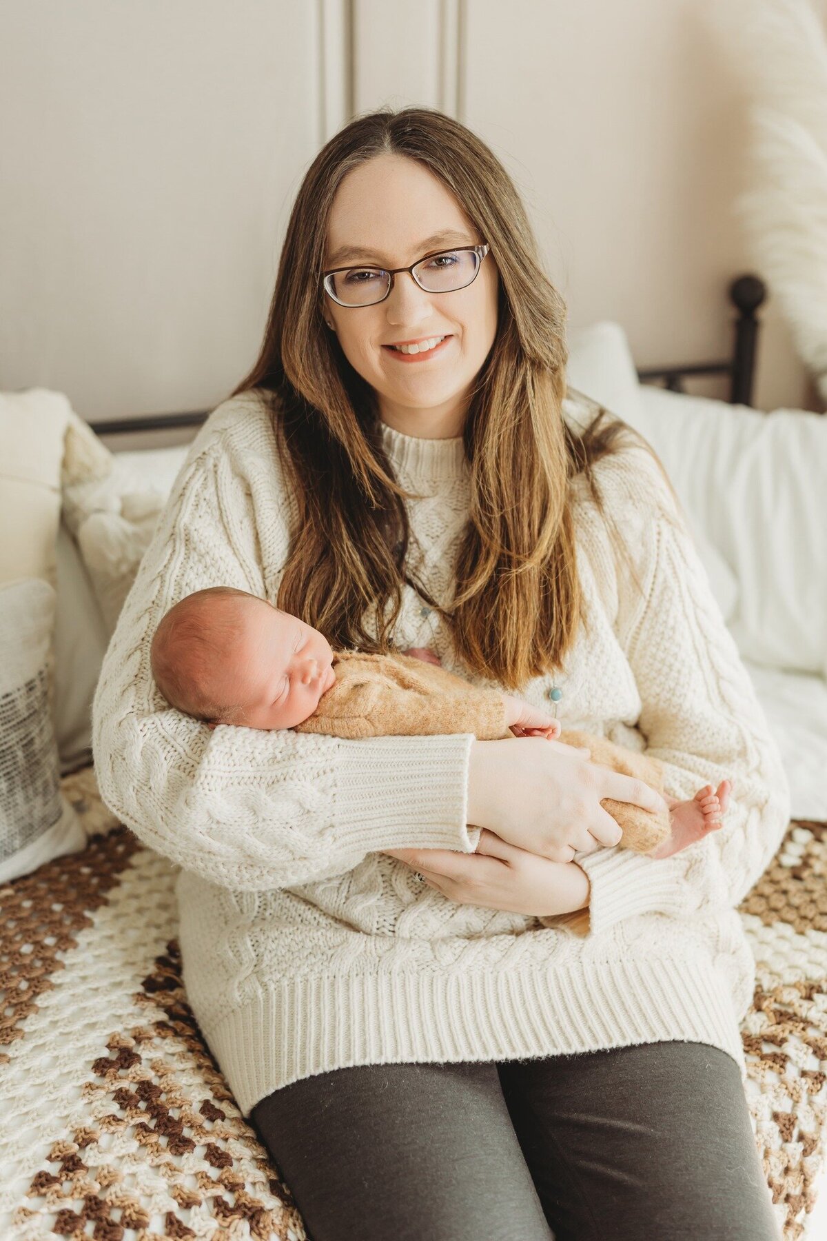 A mother with long, brown hair and glasses sits on a patterned sofa, gently cradling her newborn baby wrapped in a soft blanket. The room is softly lit, creating a warm atmosphere that conveys tenderness and joy.