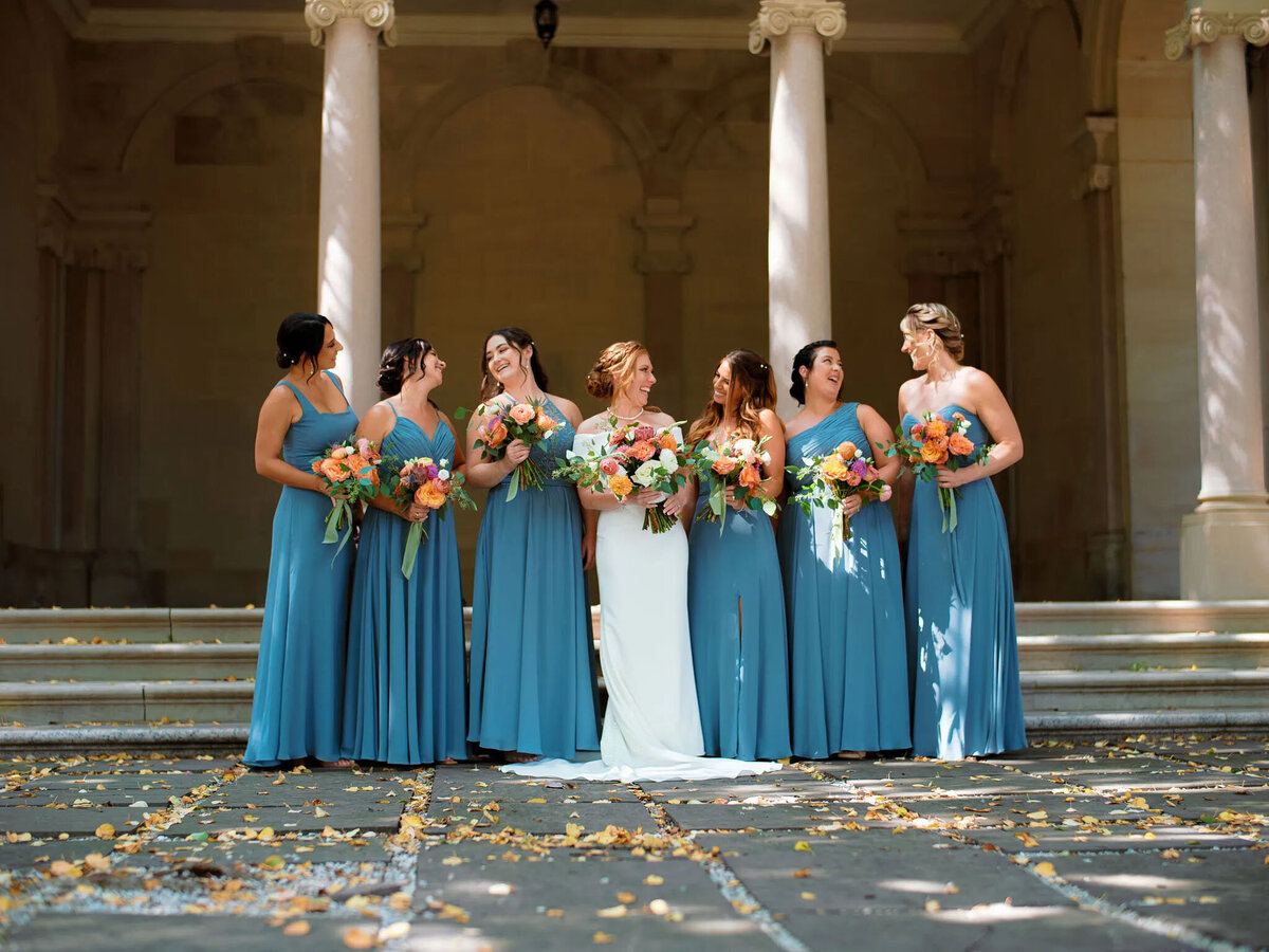 Seven women in blue dresses with bouquets smile together outdoors before stone columns; the woman in white at center suggests a bridal party, beautifully captured by an NJ wedding photographer.