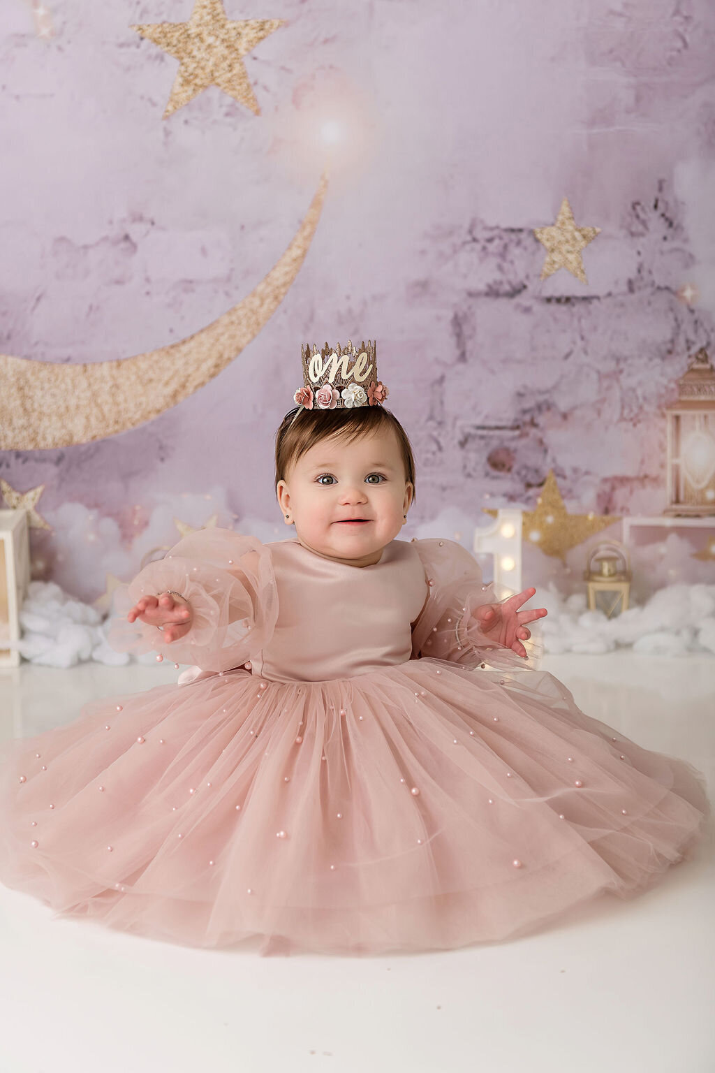 Baby girl in rose gold tulle dress celebrates first birthday milestone with hands outstretched during magical purple and gold themed portrait session