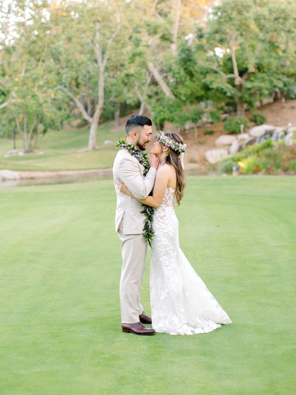 Bride and groom embracing on a green lawn, surrounded by lush trees. The bride wears a floral crown and lace gown, exuding romance and joy.