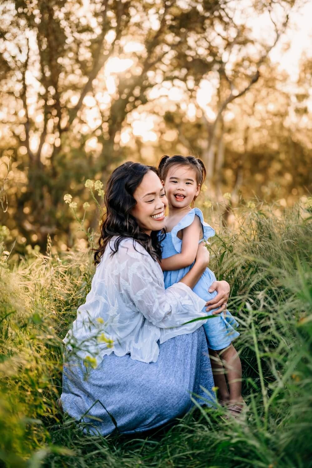 toddler looks happily at camera while mom hugs her. they are in a  lafayette wildflower field.