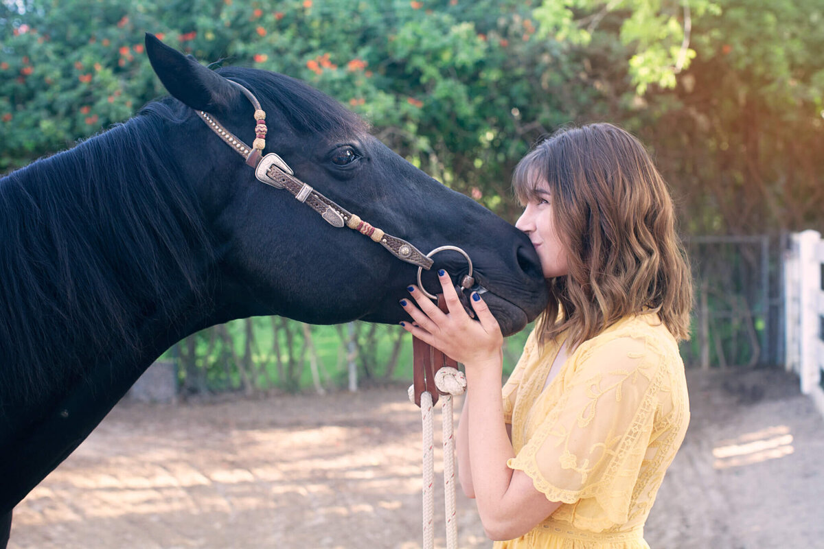 A young woman with shoulder length brown hair, wearing a yellow dress holding the chin of her black horse with a brown leash, kissing its nose and looking up into its eyes. Captured in a pasture of gravel and greenery in the background.