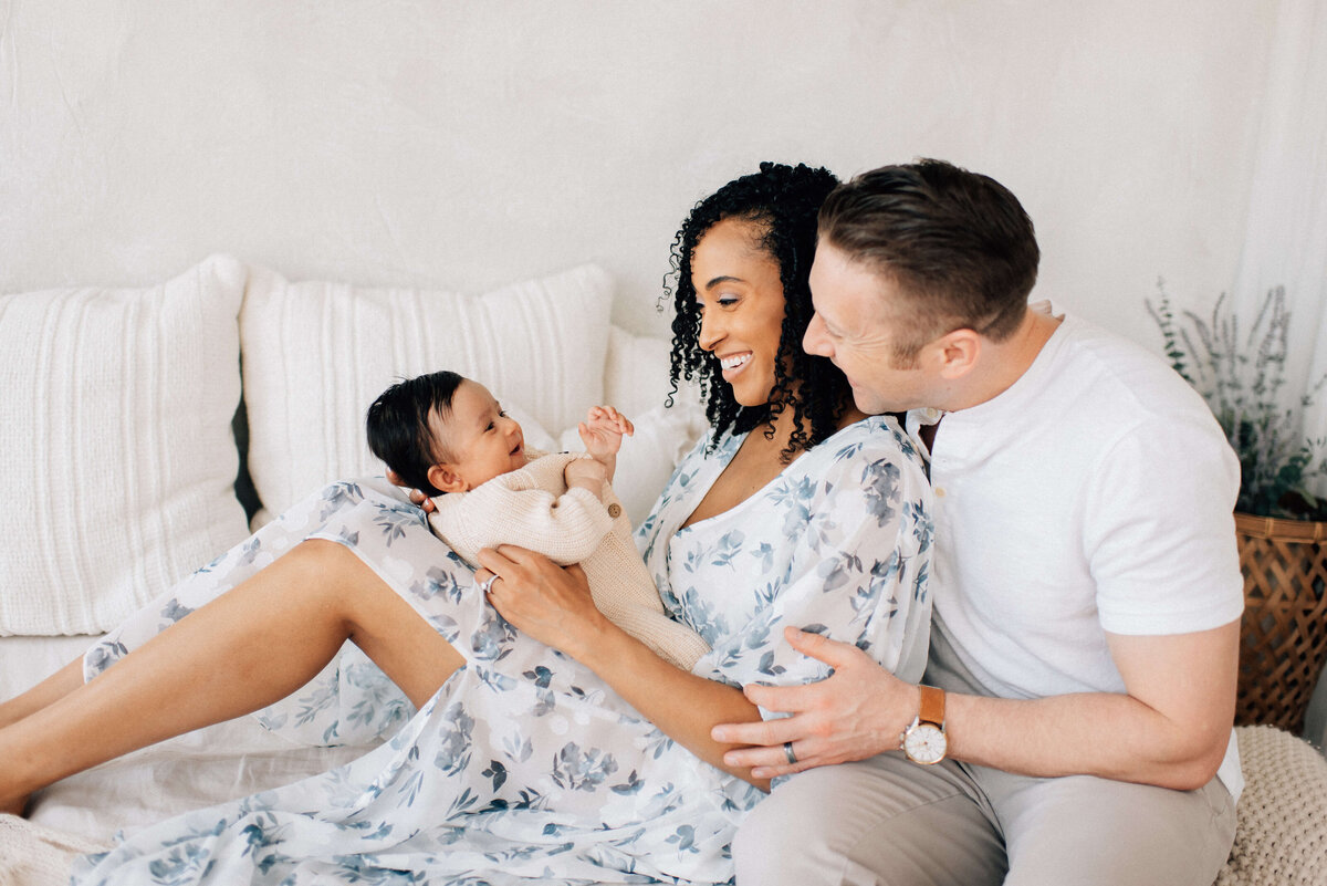 mom sitting sideways on bed with baby boy on mom's legs and dad hugging her smiling behind