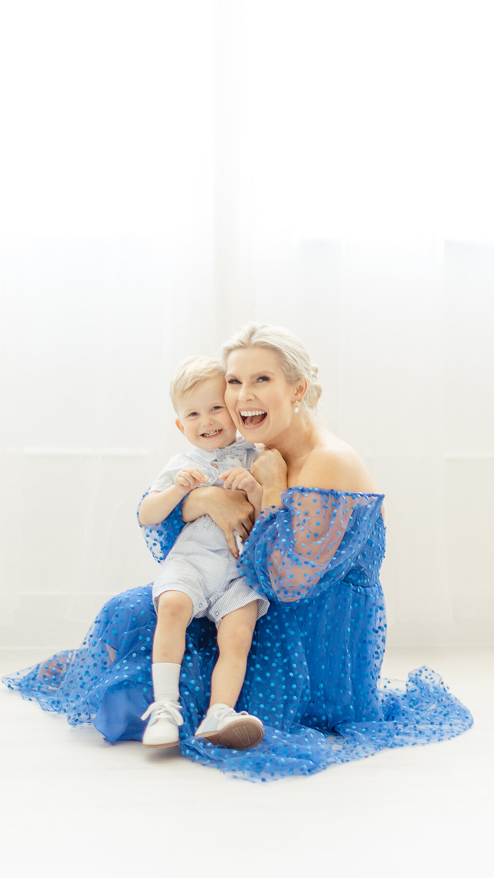 A light filled in studio motherhood photo of a mom holding her baby boy close as they look at the photographer smiling.