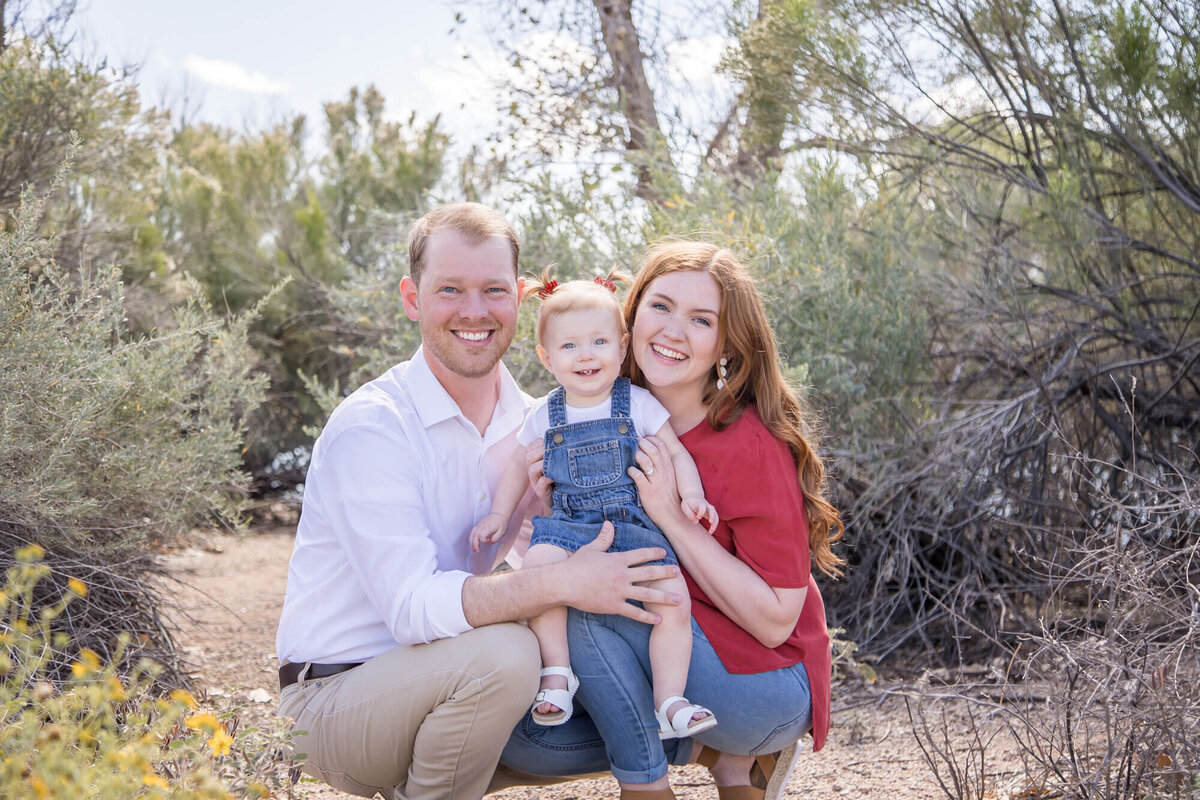 smiling Couple crouched down with toddler girl in desert park, captured by las vegas family photographer