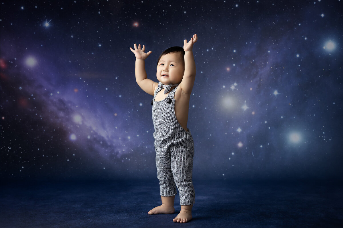 A baby in overalls and a bowtie stretches toward the stars in front of a cosmic backdrop.