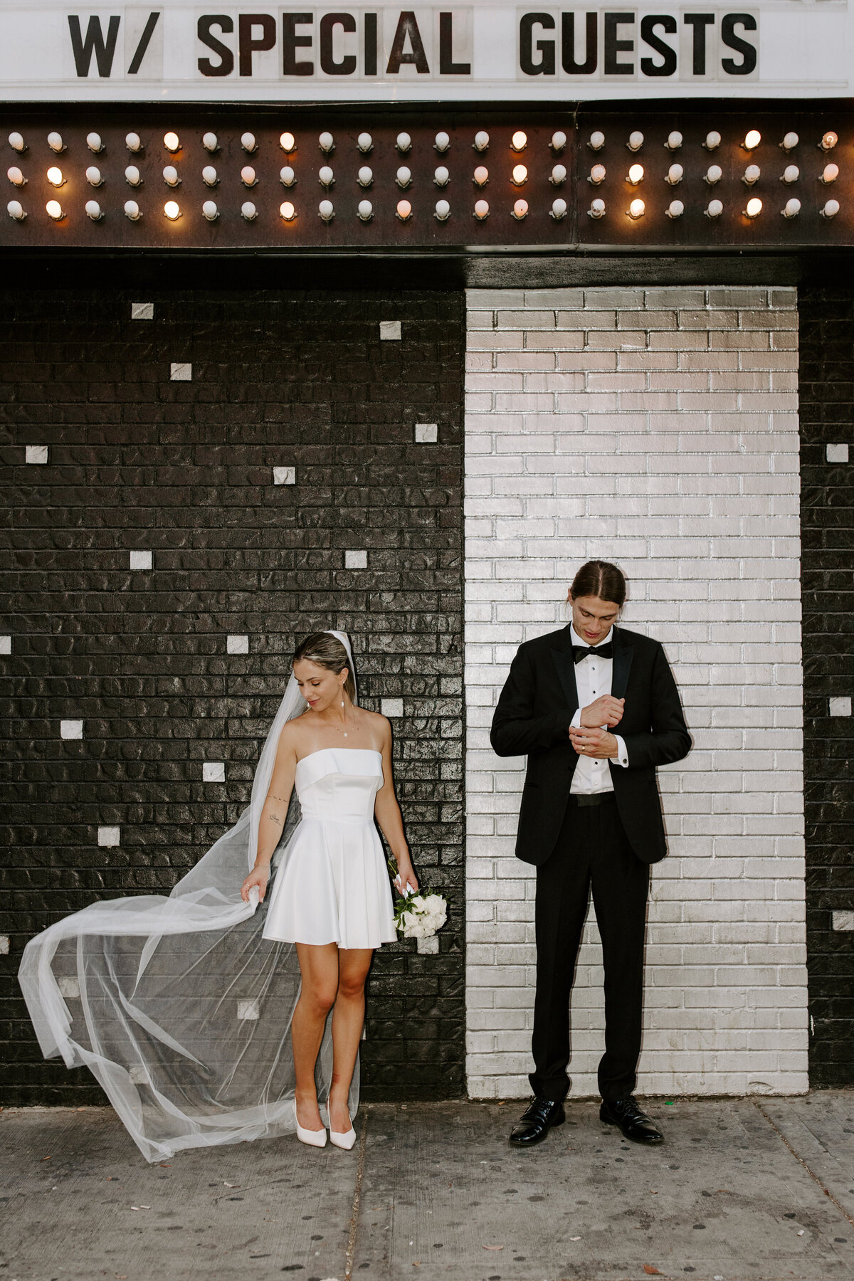 A couple poses in their elopement outfits in front of a black and white tile wall.