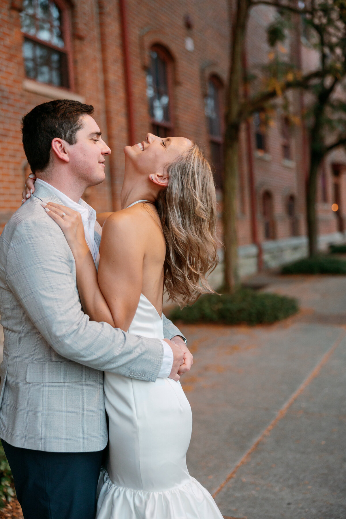 Groom embracing laughing bride in a candid moment on a sidewalk with rustic brick backdrop.