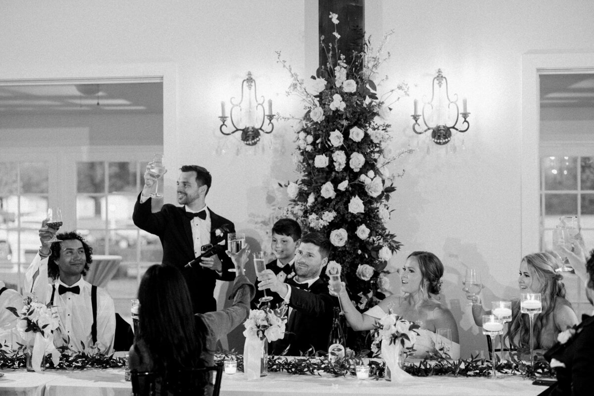 A black and white photo of people dressed in formal attire at a banquet table, with one man standing and raising a toast.