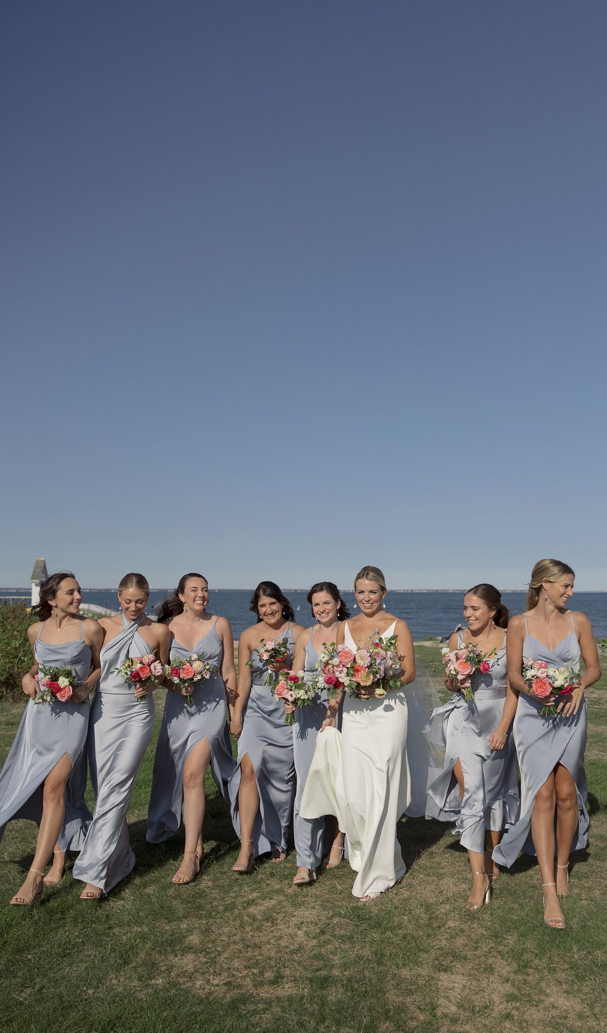 Bridesmaids walk with Bride with ocean behind them in baby blue silk dresses.