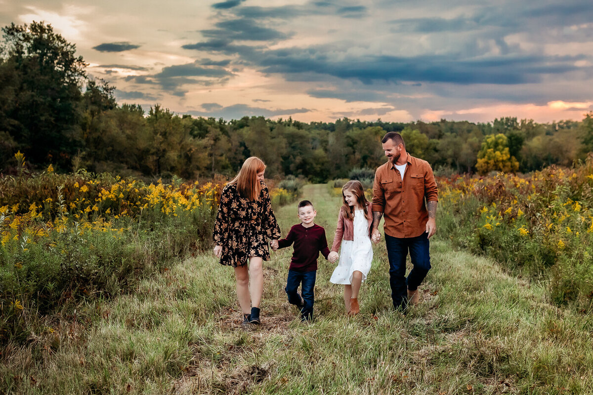 A family of four walks hand in hand along a grassy path surrounded by wildflowers. The sky is cloudy with hints of a sunset. The two children walk between the two adults, who are smiling and engaged with the children and each other.