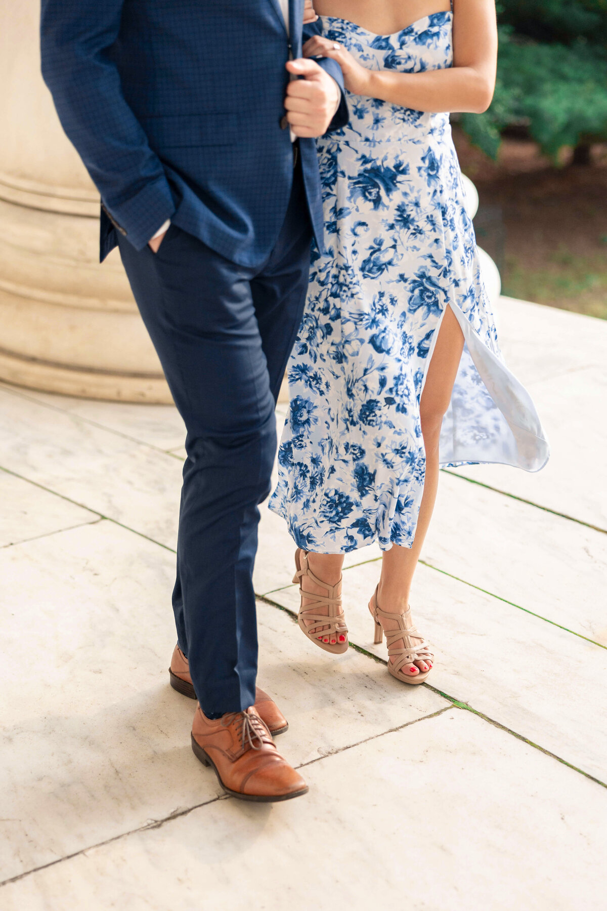 A man in a navy suit walks with a woman in a blue floral dress with a thigh-high slit. They are holding hands and walking on a stone pathway. The womans shoes are beige with heels, and the mans shoes are brown leather.