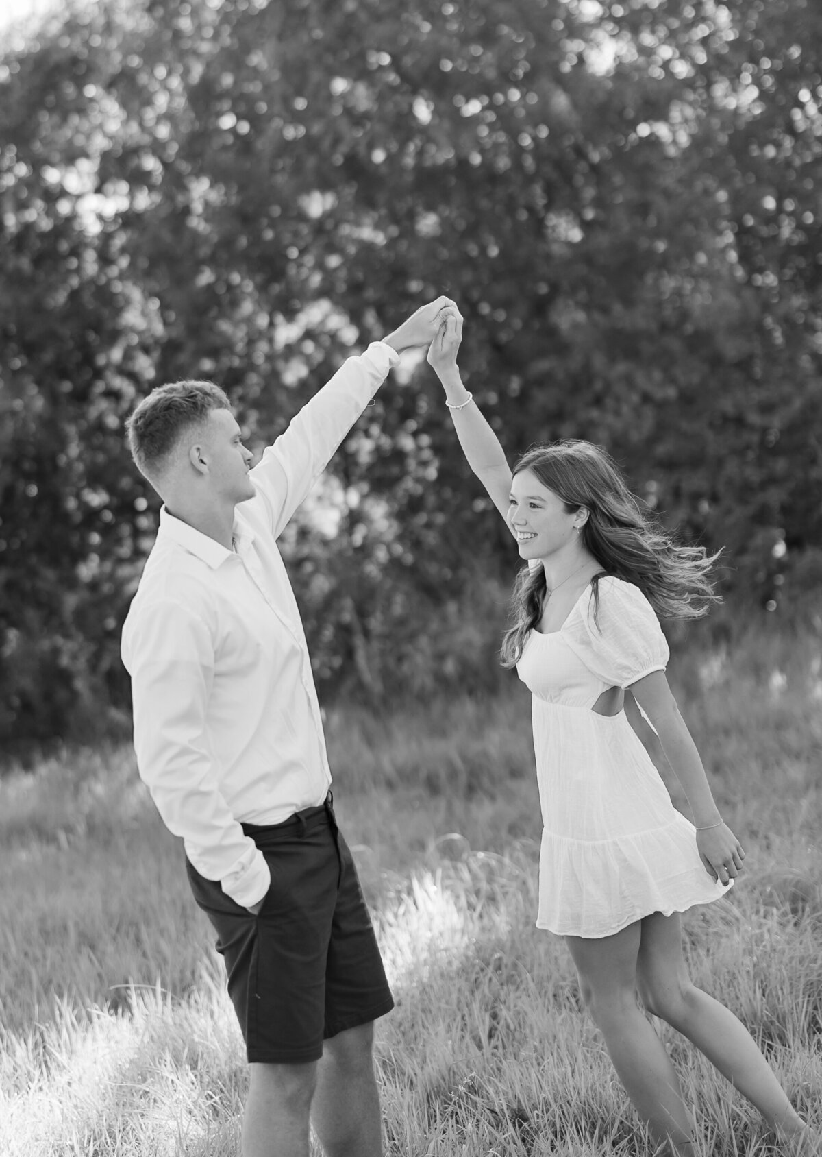 A couple dances in a sunlit grassy field. The man wears a white shirt and shorts, and the woman wears a white dress. They smile at each other with tree silhouettes blurred behind them, capturing this timeless moment as if by a skilled New England Wedding Photographer. Black and white image.