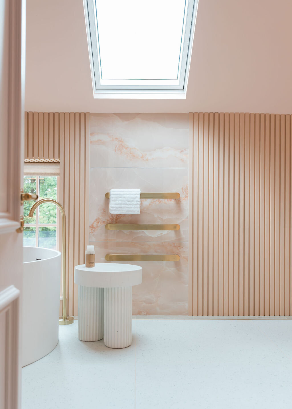 A modern bathroom featuring a round white bathtub with a sleek gold faucet. A small white table stands next to the tub. Three white towels hang on elegant gold bars mounted on a pink marble wall. A window with vertical blinds and a skylight add natural light.