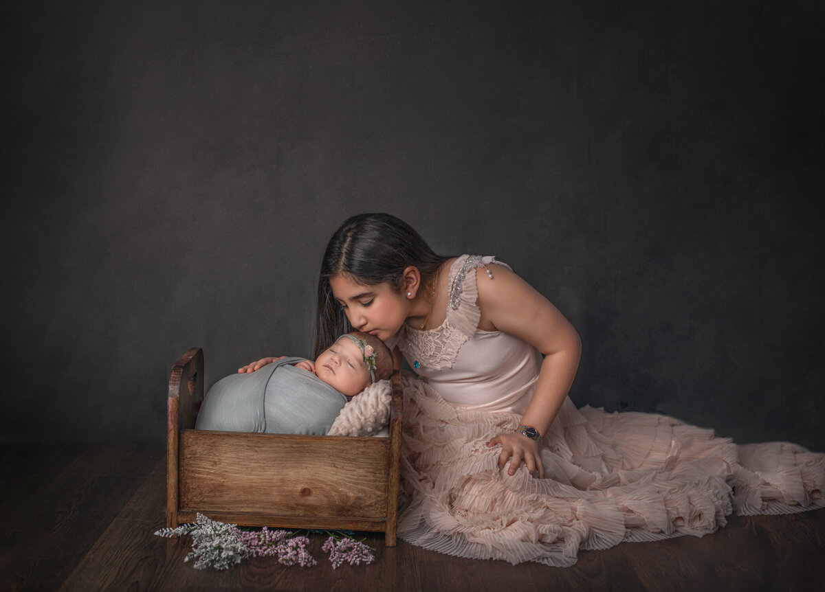 "Big sister kissing newborn baby sister lying in a wooden bed during a studio photography session in Ottawa"