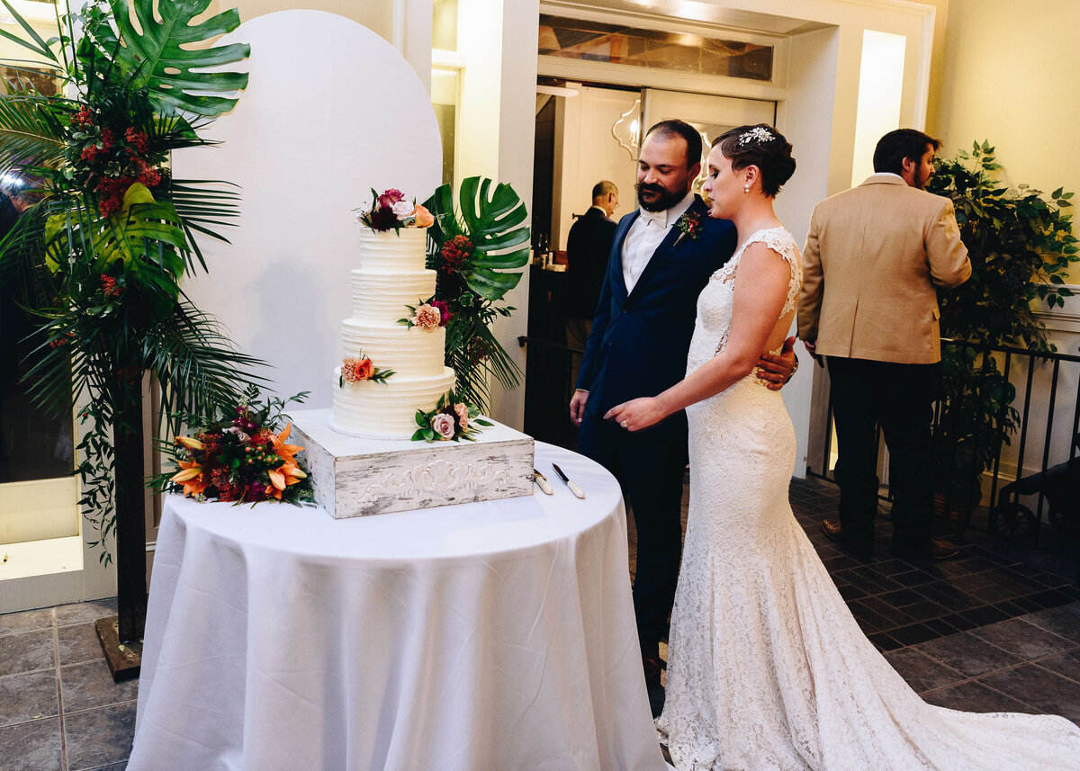 A bride and groom stand together beside a white, tiered wedding cake decorated with flowers and greenery, preparing to cut it. The cake is on a round table with a white cloth and floral arrangements nearby.