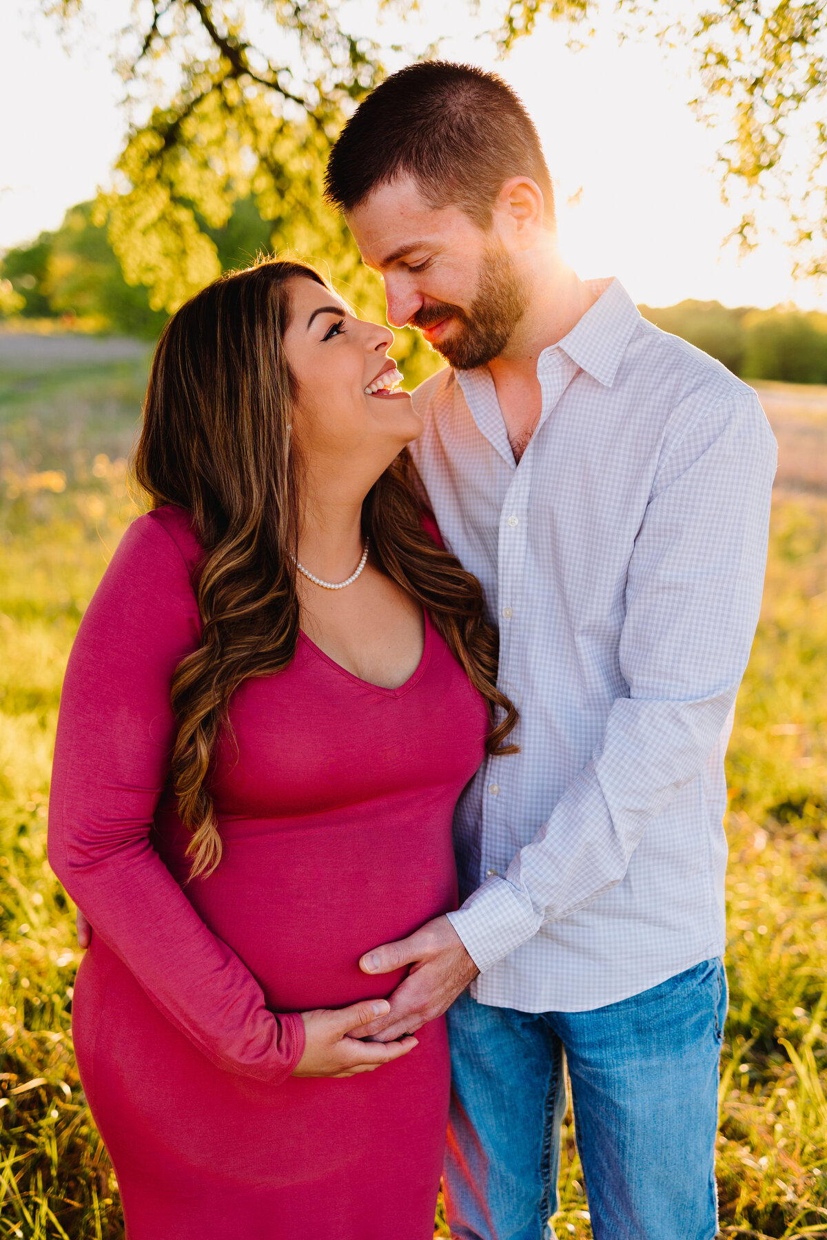 Maternity photo of a pregnant couple in a garden. The woman, dressed in a flowing long pink dress, smiles affectionately at the man. He gently touches her belly, dressed in blue jeans and a blue shirt.