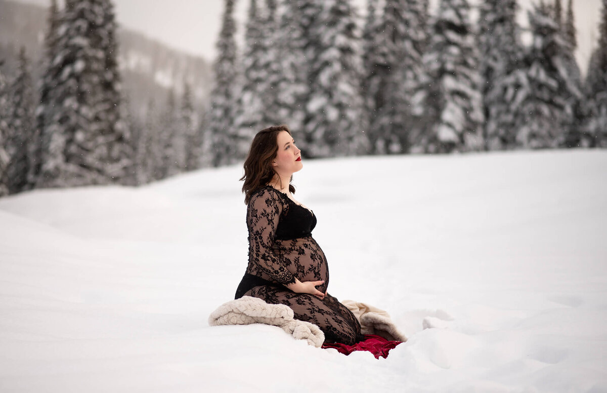 In sun peaks, a pregnant women sits on a blanket in the snow while holding her belly and looking off into the distance. Taken by Brittany Danielle Photography