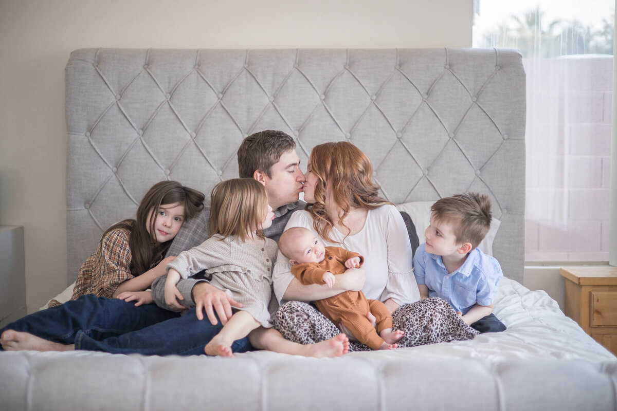 Couple kissin g surrounded by their four small children on a bed