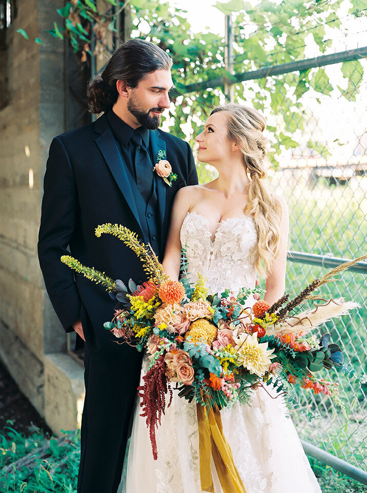 Bride and groom, wearing a black tuxedo and white wedding gown,  make eye contact with a large bouquet of flowers in hand.