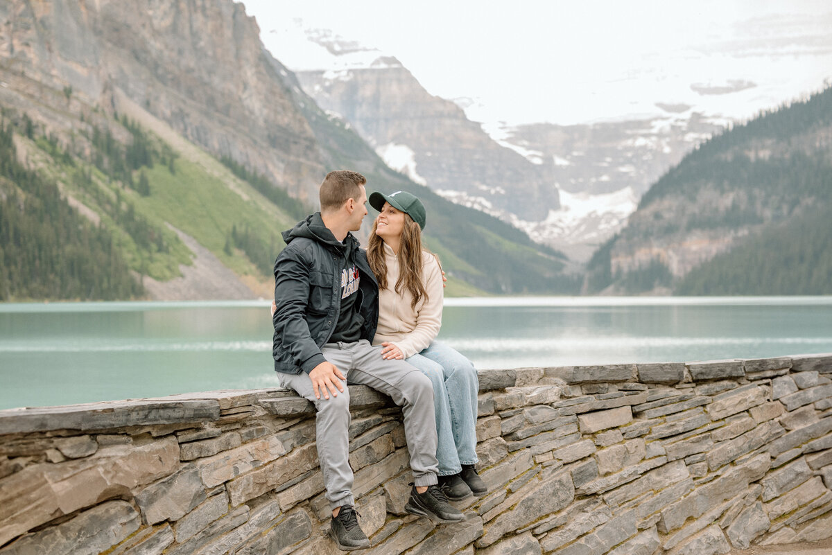 Engaged couple sitting on stone ledge at Lake Louise with peaceful water and sky.