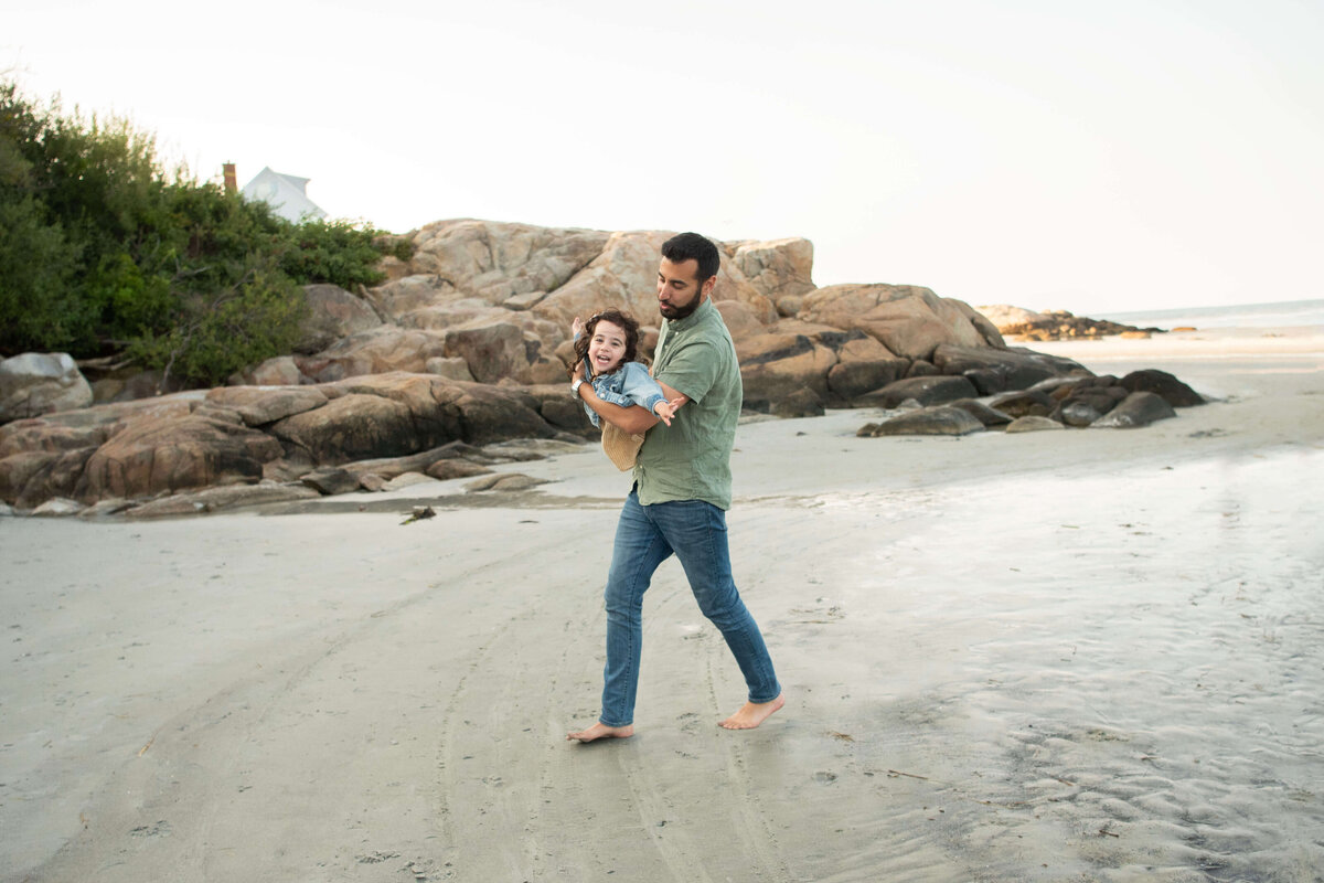 A dad runs and holds his daughter with her arms out at Wingaersheek beach.