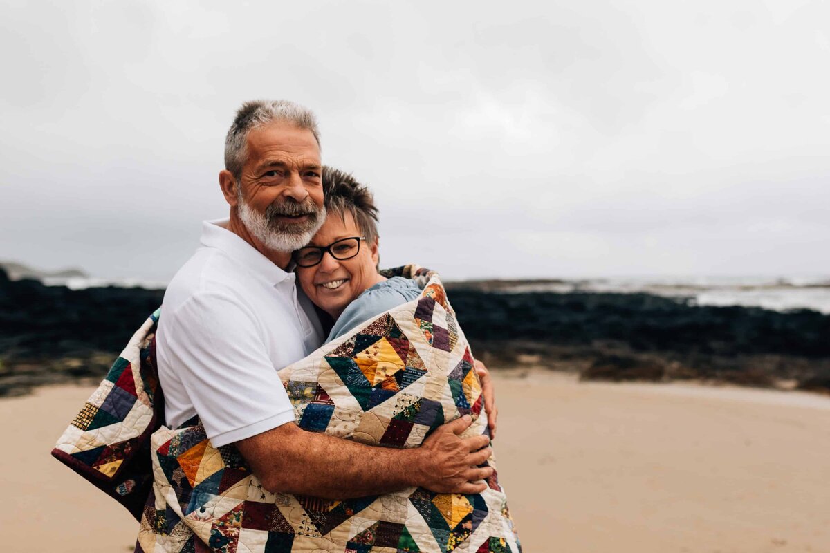 Woman wrapped in quilt with man hugging her, at beach during couple photoshoot Melbourne, And So I Don't Forget Photography