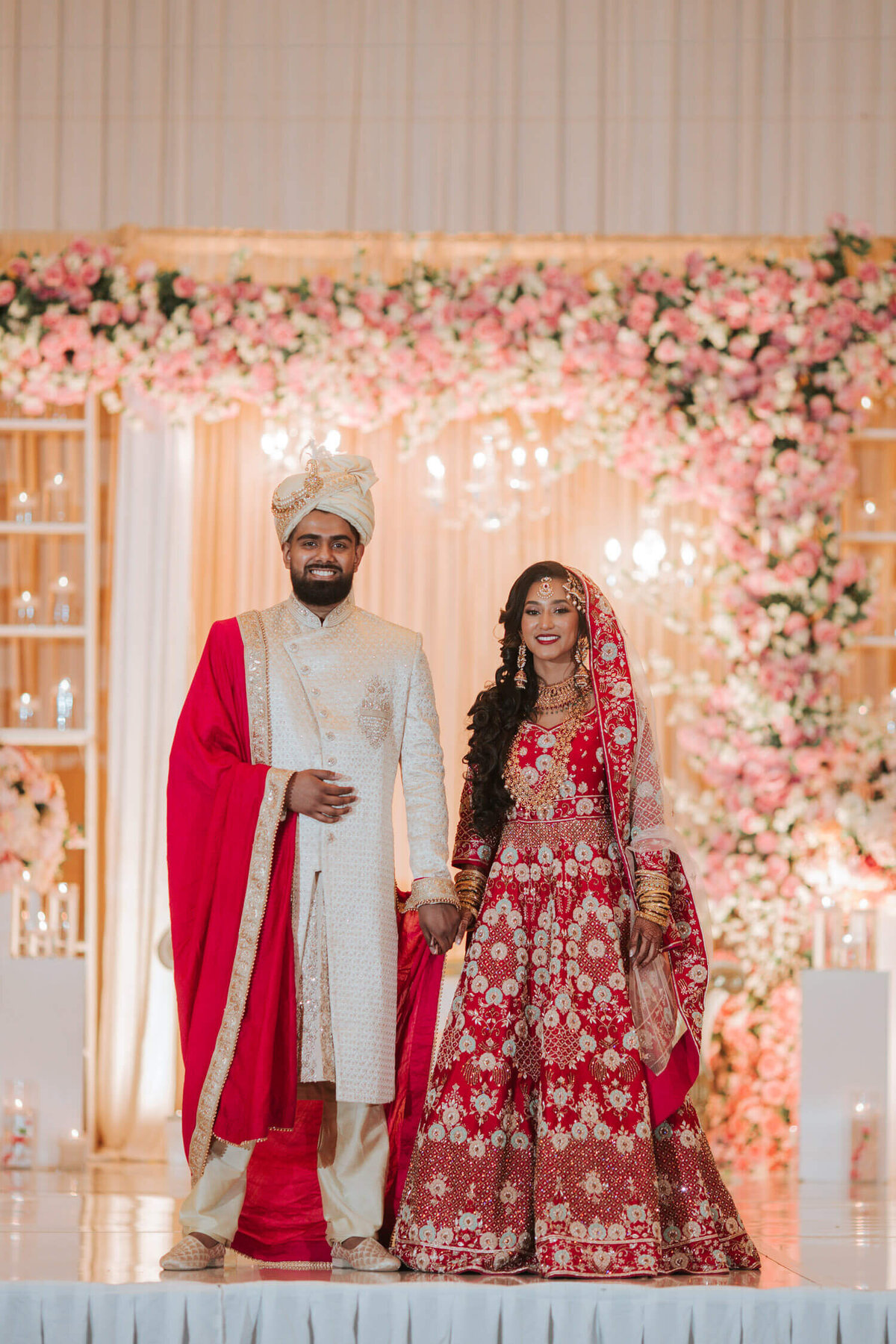 A couple dressed in elegant red and white traditional wedding attire stands before a floral backdrop at Royal Albert's Palace.