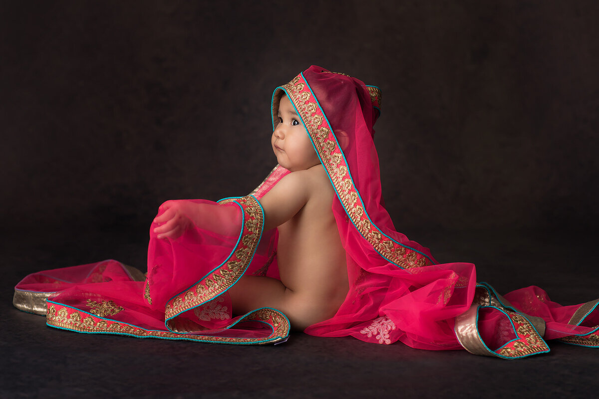 A baby sits wrapped in a vibrant fuchsia and gold sari with turquoise trim, gazing off to the side against a dark backdrop, highlighting a luxurious Dallas milestone photography session with cultural elegance.