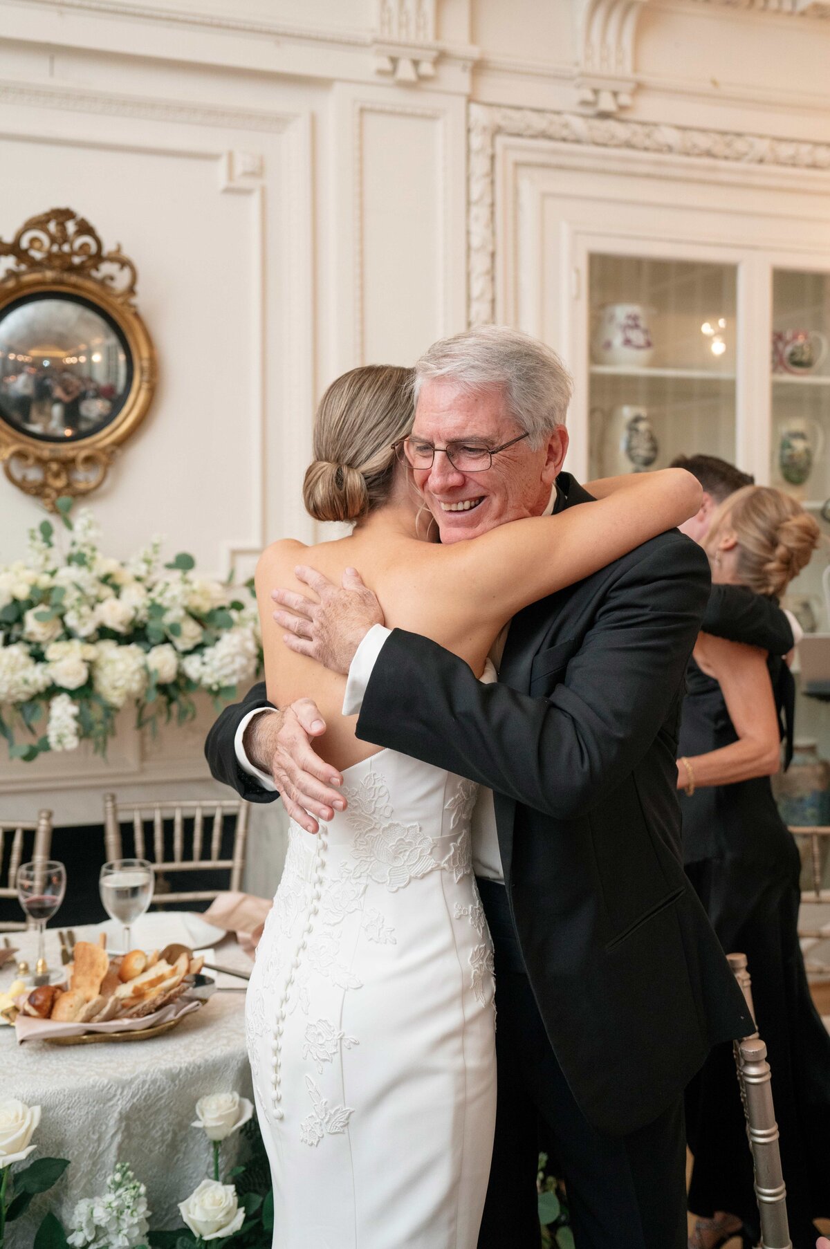 A bride in a white dress hugs an older man in a suit inside an elegantly decorated room. Theres a table set with bread and wine, and floral arrangements in the background. Both appear joyful and emotional.